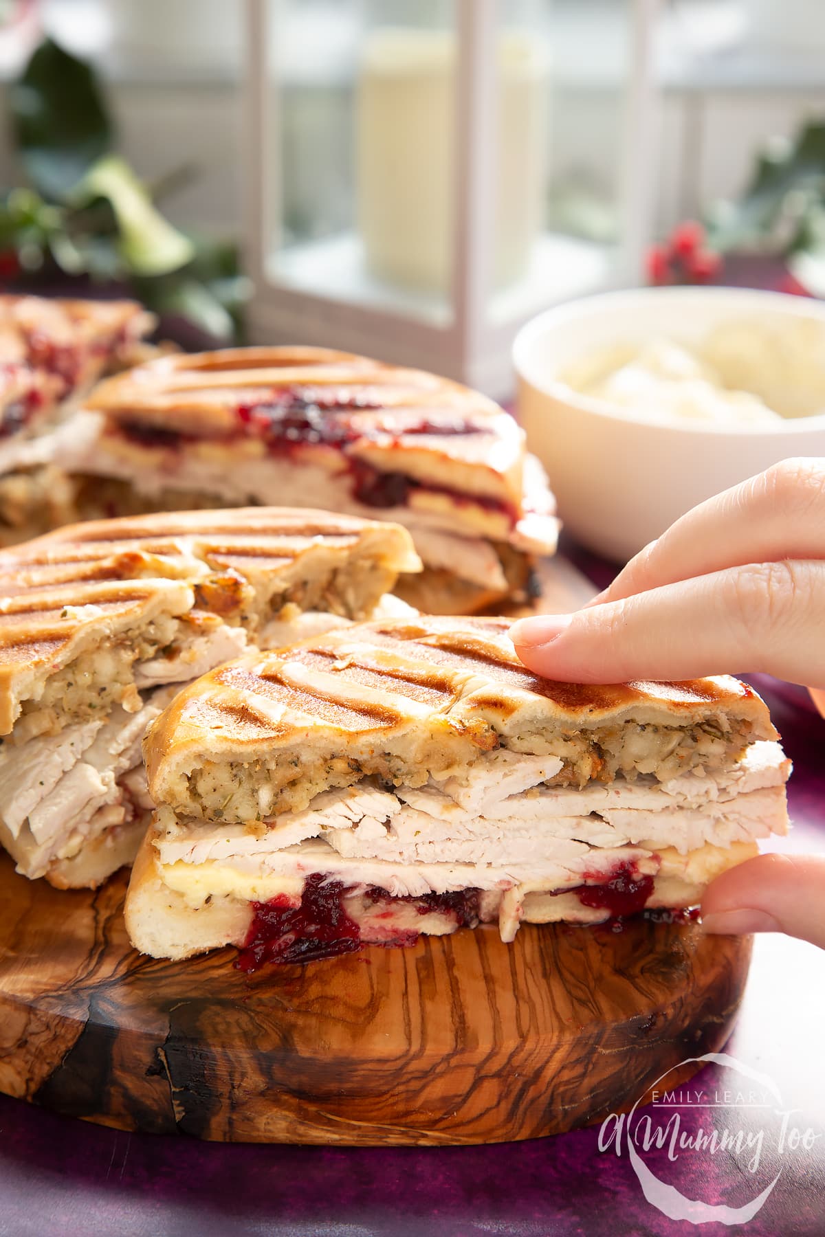 Front angle shot of a hand holding a Toasted turkey bagel served on a wooden plate with a mummy too logo in the lower-right corner