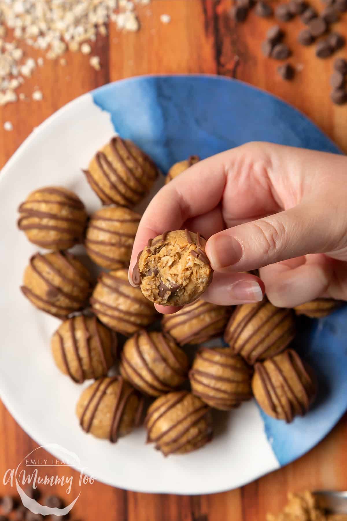 3 ingredient peanut butter and oatmeal balls decorated with chocolate on a blue and white plate. A hand holds one that has a bite out of it.