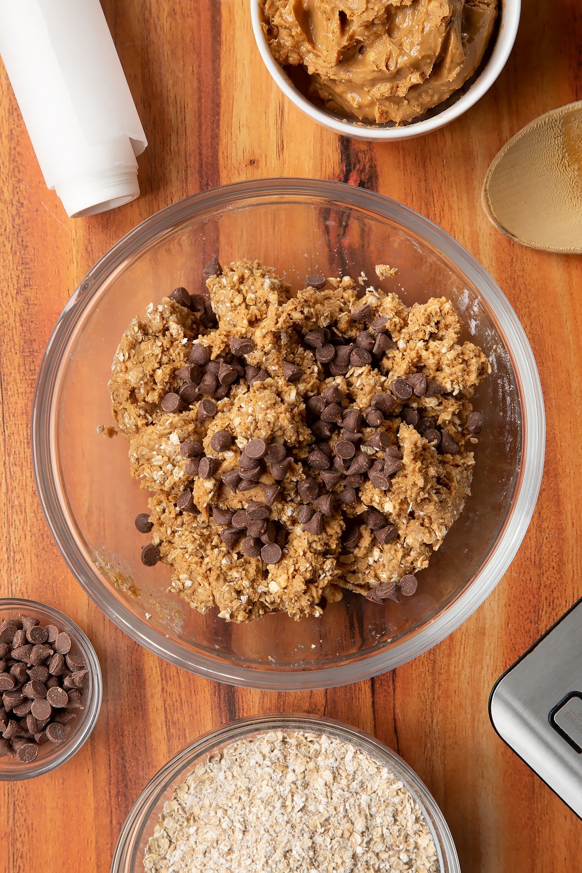 Rolled oats and peanut butter mixed together in a glass mixing bowl with chocolate chips on top. Ingredients to make 3 ingredient peanut butter and oatmeal balls surround the bowl.