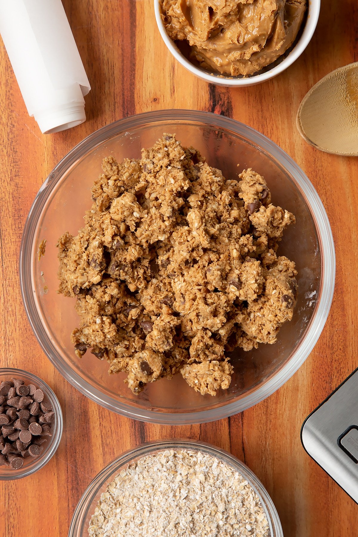 Rolled oats, peanut butter and chocolate chips mixed together in a glass mixing bowl. Ingredients to make 3 ingredient peanut butter and oatmeal balls surround the bowl.