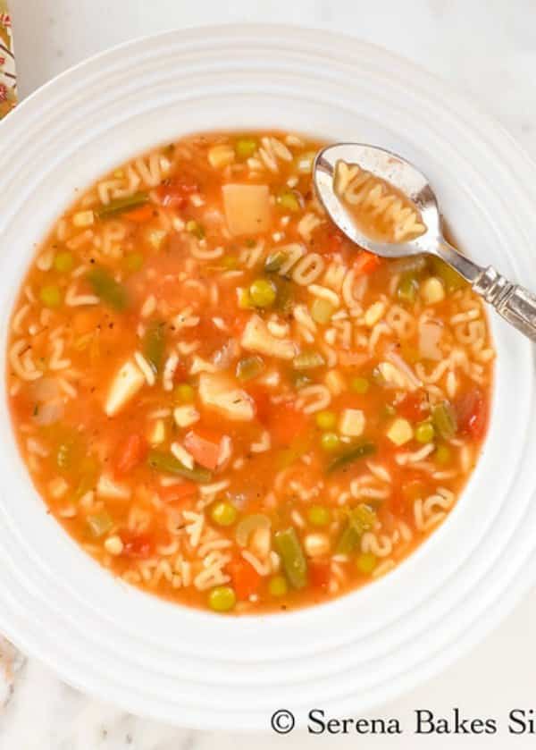 A decorative silver spoon with the alphabet soup spelling 'yummy'. In the background sits the remainder of the bowl of soup inside a white bowl.