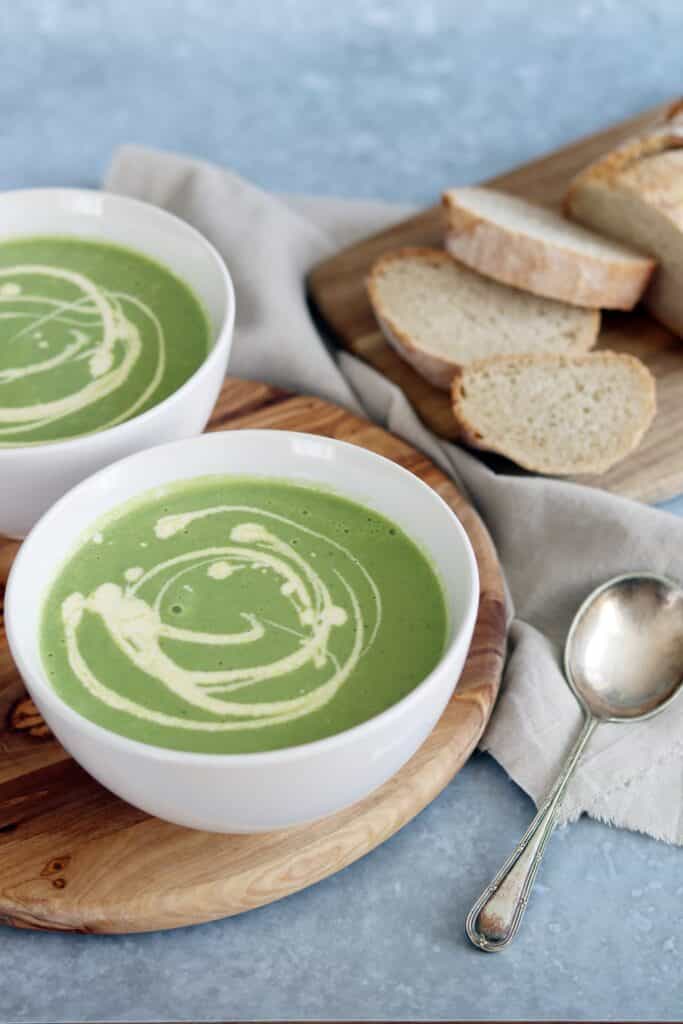 Two bowls of broccoli soup sit on a wooden lazy susan. At the side there's an organic table cloth and a vintage silver spoon.