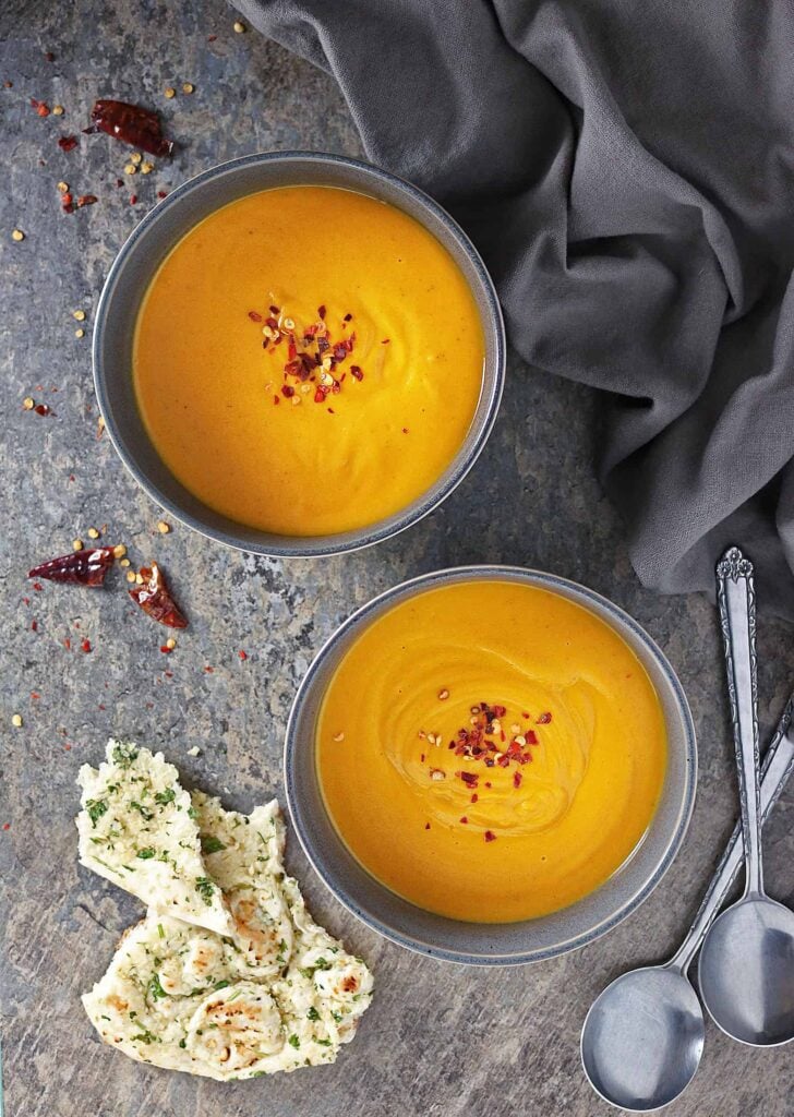 A grey bowls filled with Roasted Sweet Potato & Ginger Soup  on a dark grey background. The sides are decorated with spoons and chilli flakes.