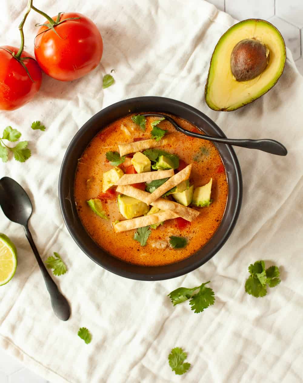 Chicken Tortilla Soup in a dark bowl with a black spoon. The bowl is on a white table cloth and surrounded by tomatoes, fresh herbs and avocado.