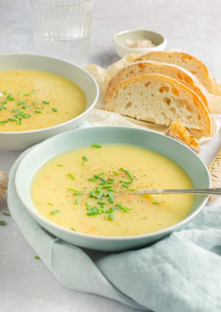 Two bowls of leek and potato soup sit on top of a silver table top surrounded by chunks of bread roll.