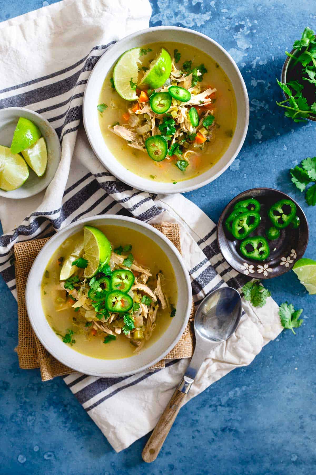 Bowls of Spicy Chicken Lime Soup with bowls of chopped peppers, a tea towel, limes and a spoon.