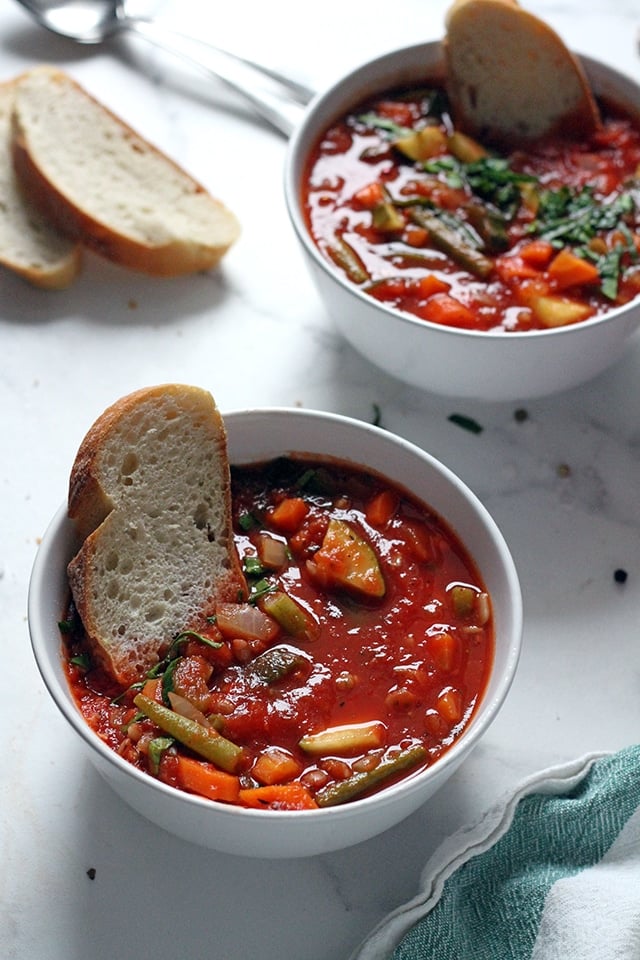 A white bowl filled with Minestrone Soup and a slice of crusty bread on a white marble tabletop.