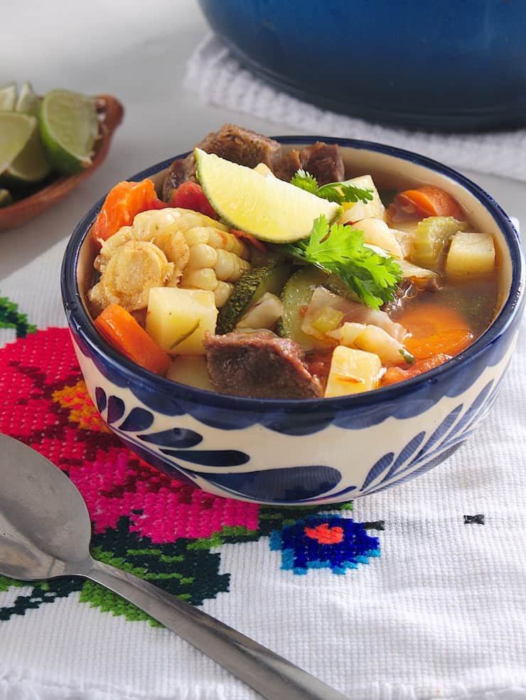 Blue and white decorative bowl filled with Caldo de Res (Beef Soup). In the corner there's a plate of cut up limes and a colourful napkin.