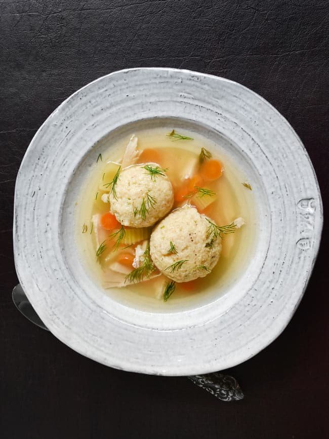 A rustic bowl filled with Matzo Ball Soup sits on a wooden tabletop.