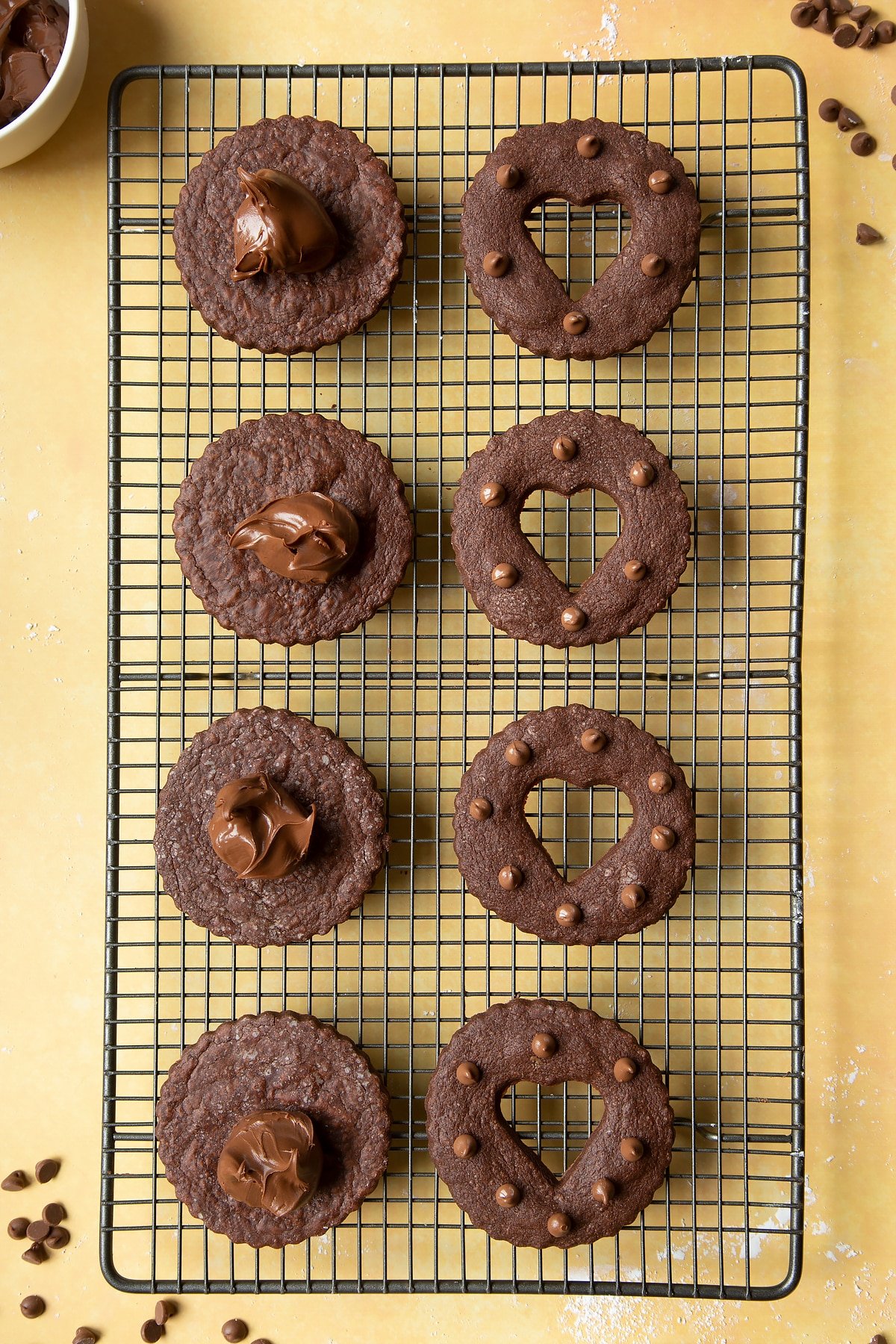 Nutella sandwich cookies on a cooling rack ready to assemble. The bottom halves have a dollop of Nutella on each of them.