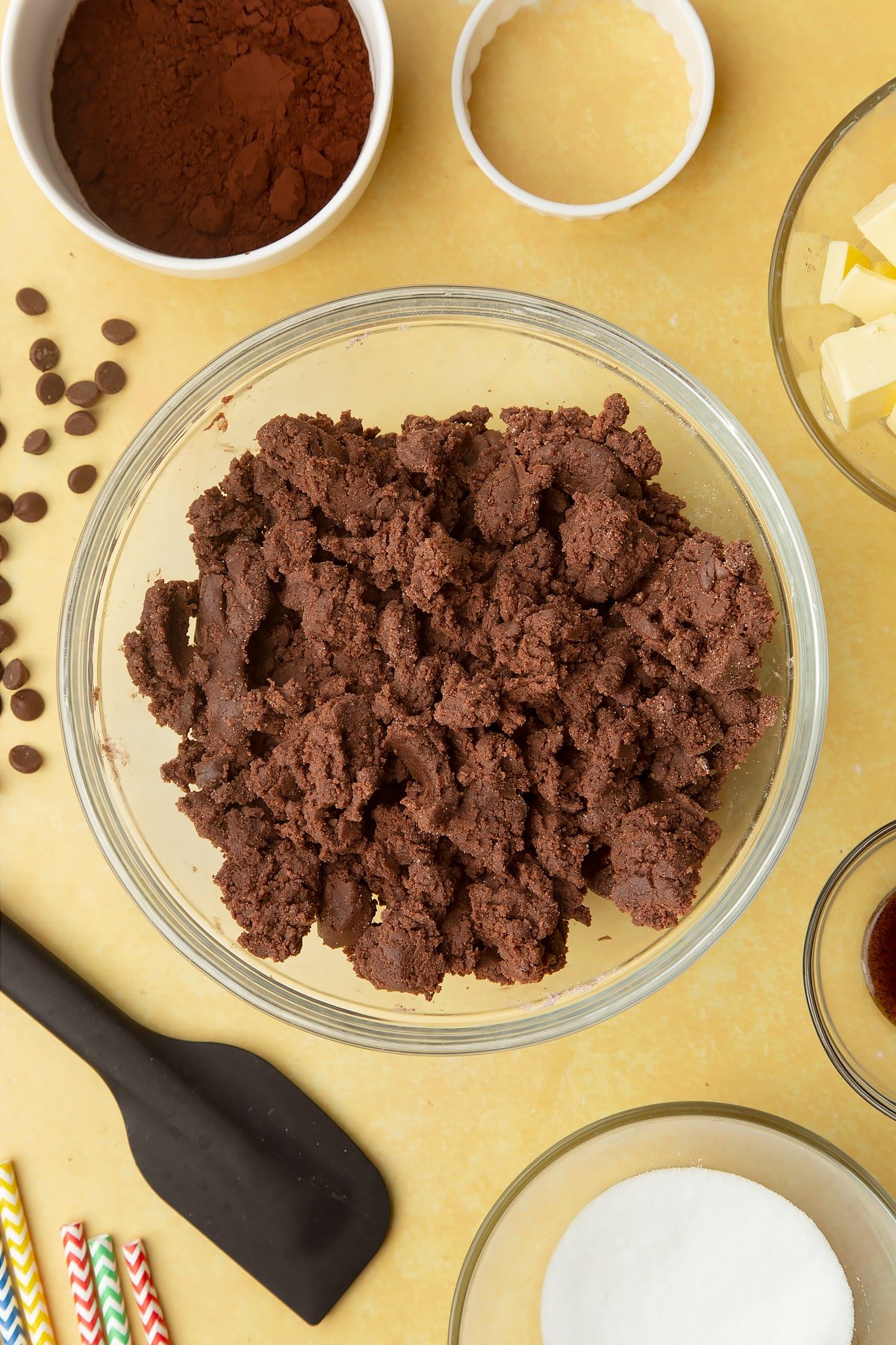 A bowl of roughly mixed chocolate shortbread dough. Ingredients to make Nutella sandwich cookies surround the bowl.