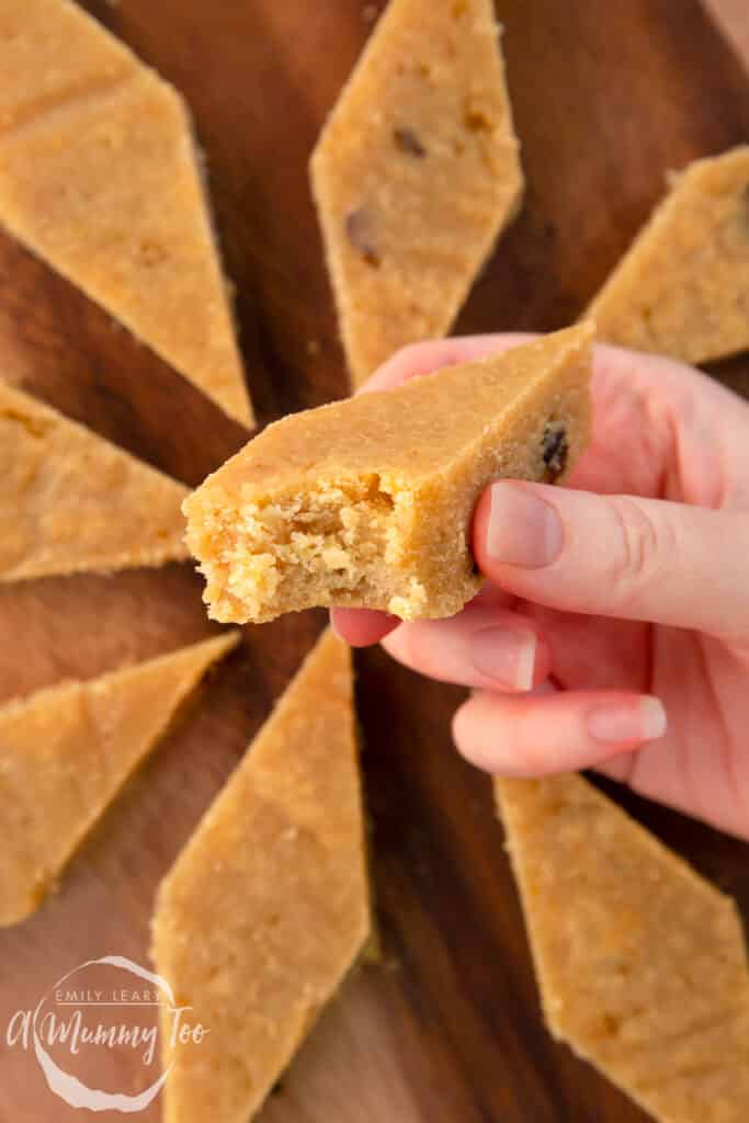 Hand holding a piece of sanwin-makin (golden semolina cake) with a bite out of it. More is shown on a wooden board.