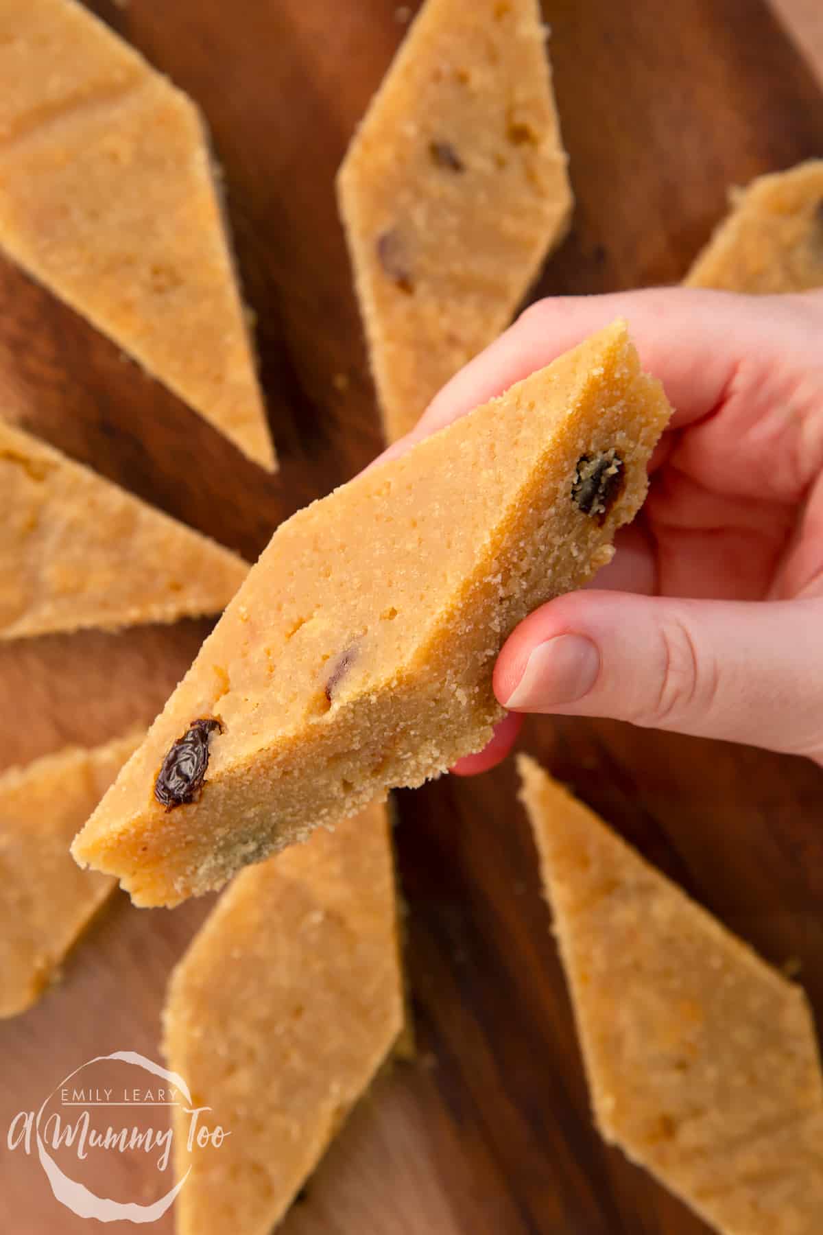 Hand holding a piece of sanwin-makin (golden semolina cake). More is shown on a wooden board.