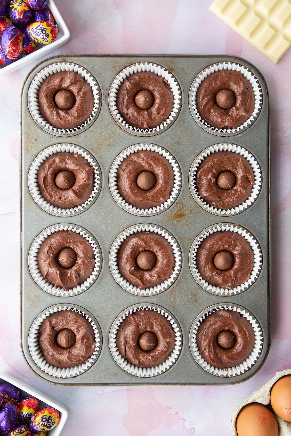 Chocolate cake batter in a 12 hole muffin tray with a Creme Egg Mini pushed into the centre of each one. Ingredients to make Cadbury Creme Egg cakes surround the tray.