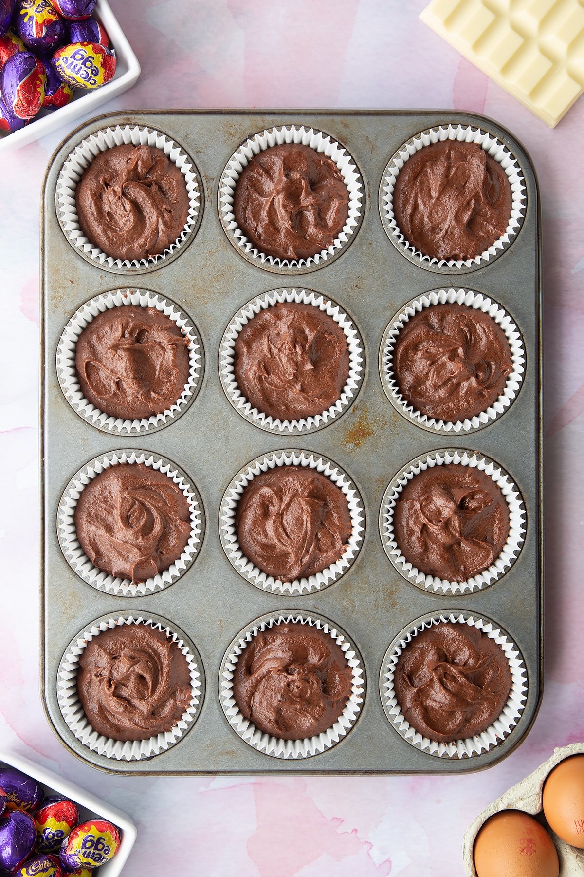 Chocolate cake batter in a 12 hole muffin tray with a Creme Egg Mini hidden in the centre of each one. Ingredients to make Cadbury Creme Egg cakes surround the tray.