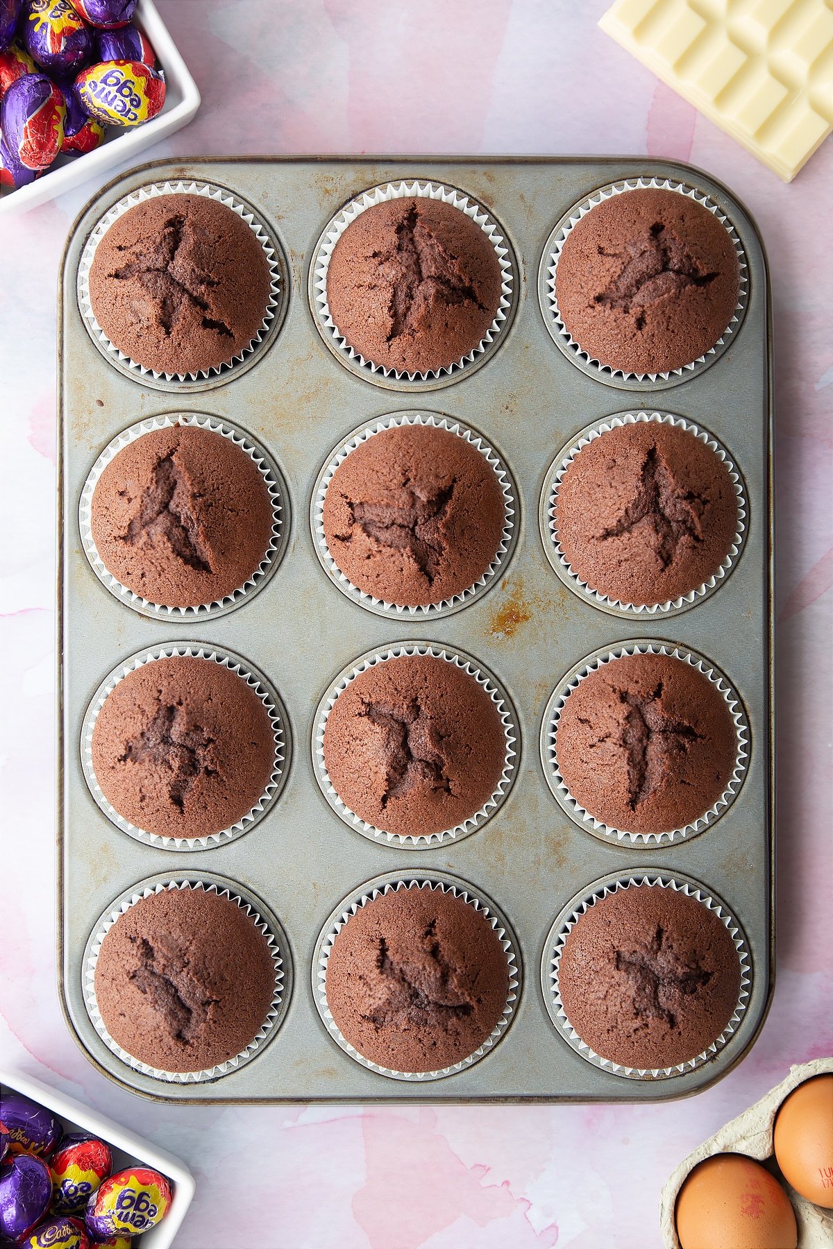 Chocolate cupcakes in a 12 hole muffin tray. Ingredients to make Cadbury Creme Egg cakes surround the tray.