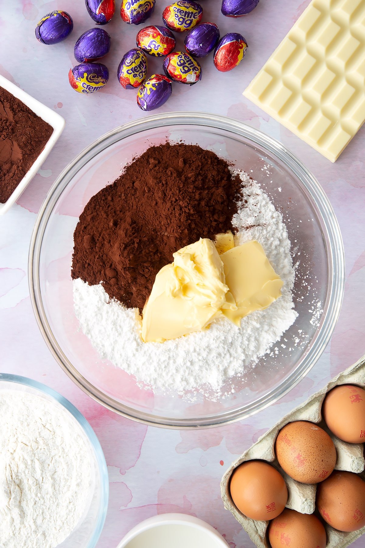 Butter, icing sugar, cocoa powder and water in a bowl. Ingredients to make Cadbury Creme Egg cakes surround the bowl.