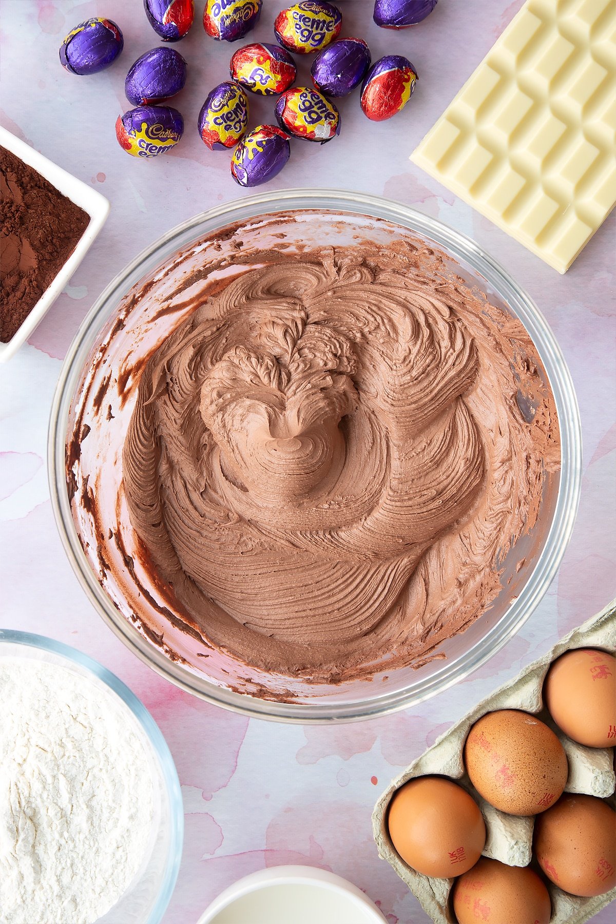 Chocolate frosting in a bowl. Ingredients to make Cadbury Creme Egg cakes surround the bowl.