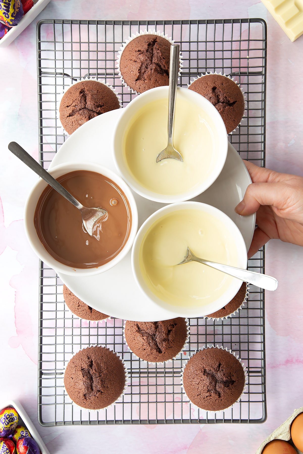 Three bowls of melted chocolate, two white and one milk. Below are chocolate cupcakes on a cooling rack.