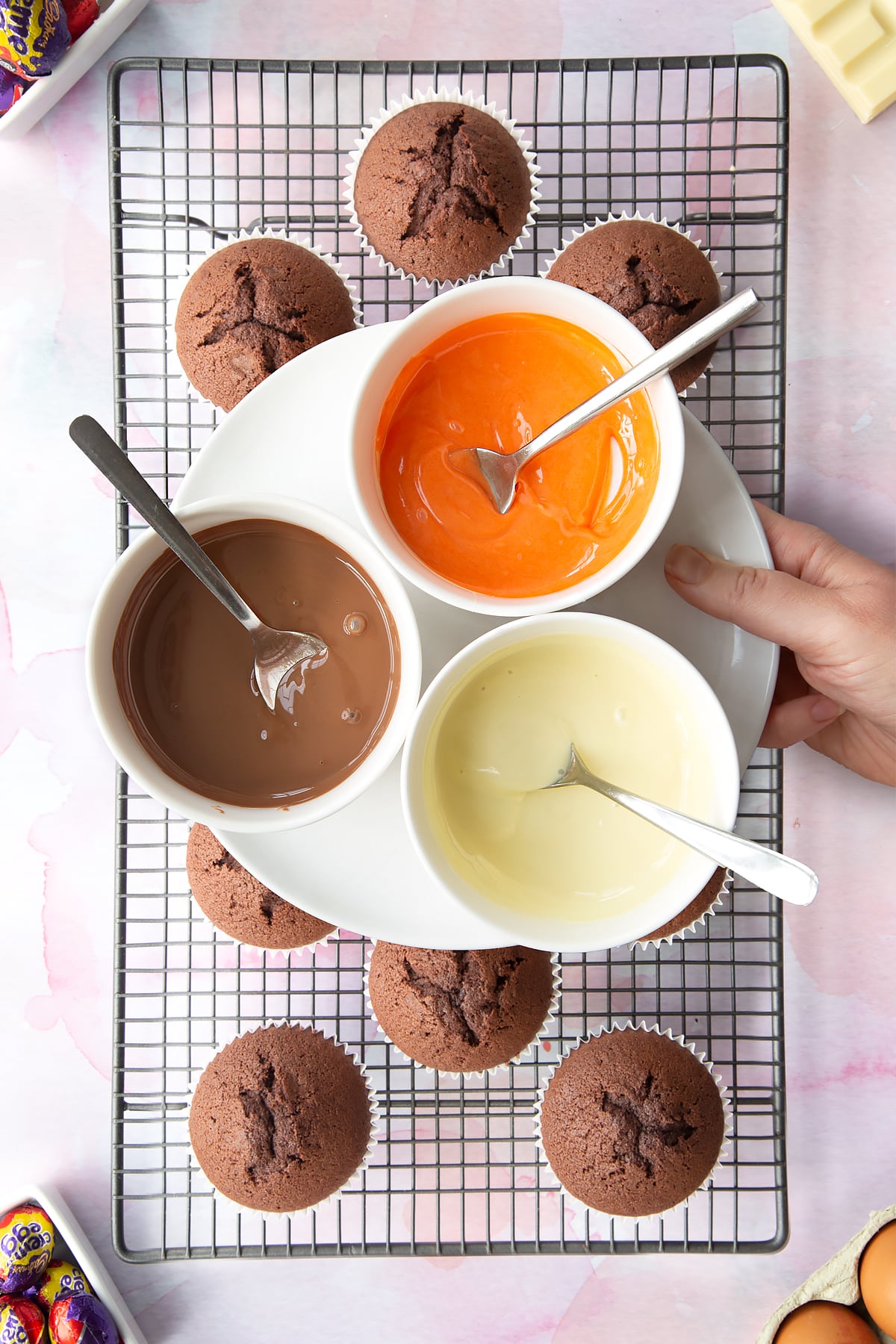 Three bowls of melted chocolate, one white, one orange and one milk. Below are chocolate cupcakes on a cooling rack.