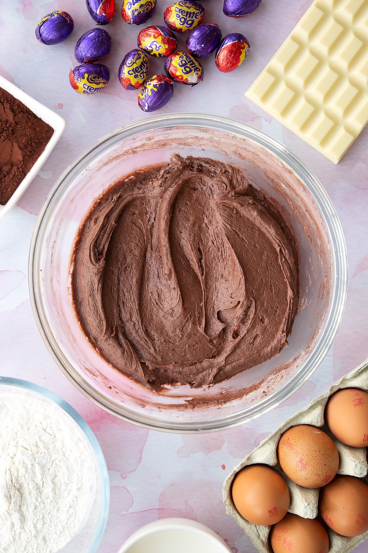 Thick chocolate cake batter in a bowl. Ingredients to make Cadbury Creme Egg cakes surround the bowl.