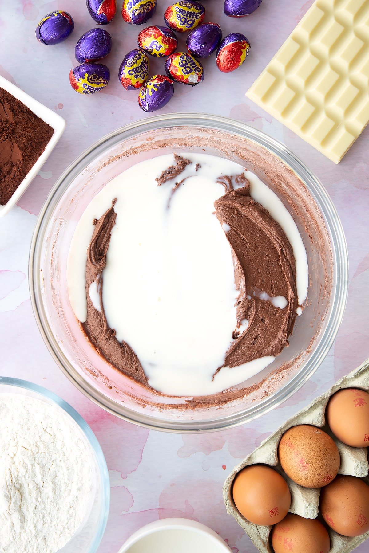 Thick chocolate cake batter in a bowl with milk on top. Ingredients to make Cadbury Creme Egg cakes surround the bowl.