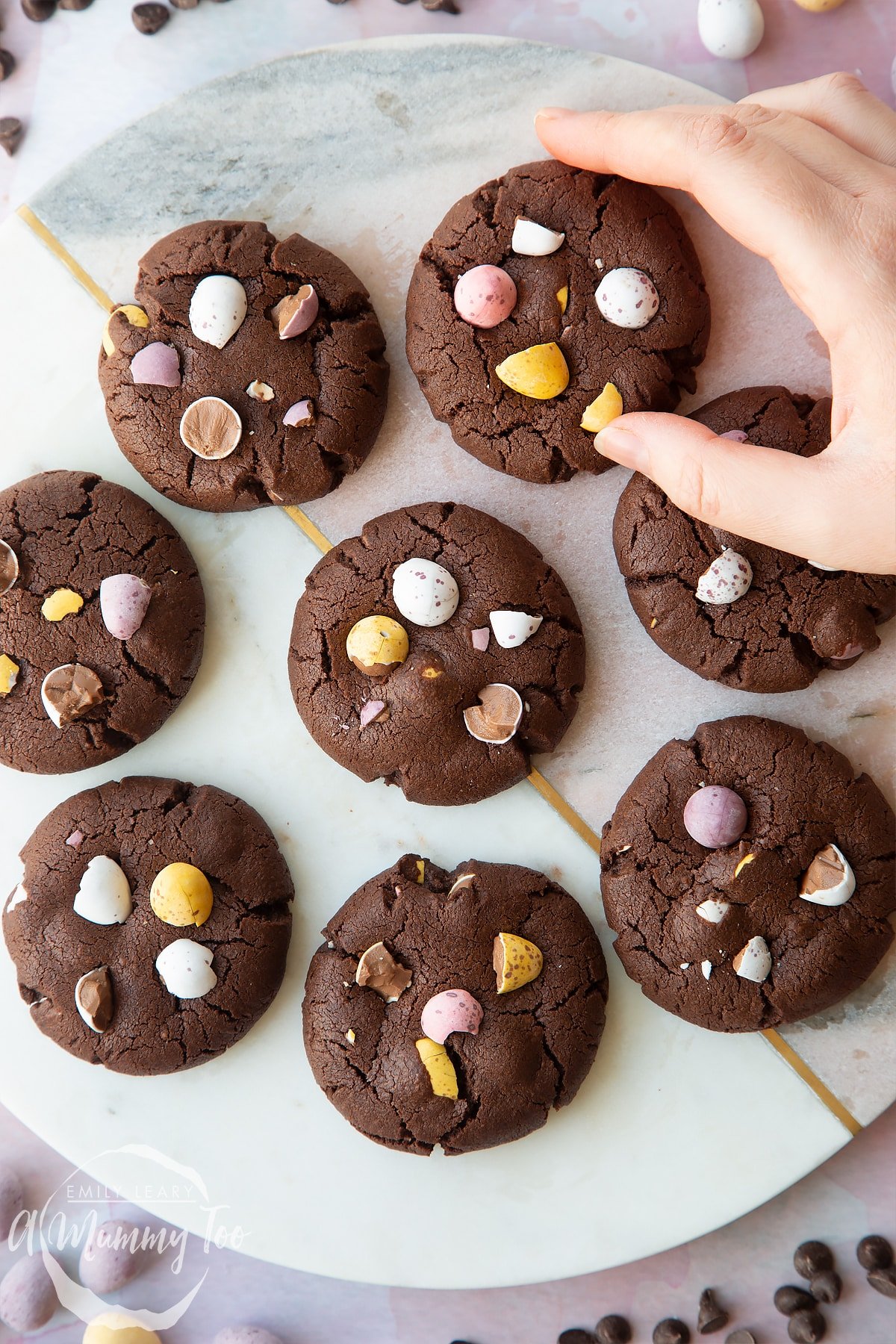 Chocolate Easter cookies arranged on a round marble board. The cookies are chocolate brown and topped with Mini Eggs. A hand reaches for one.