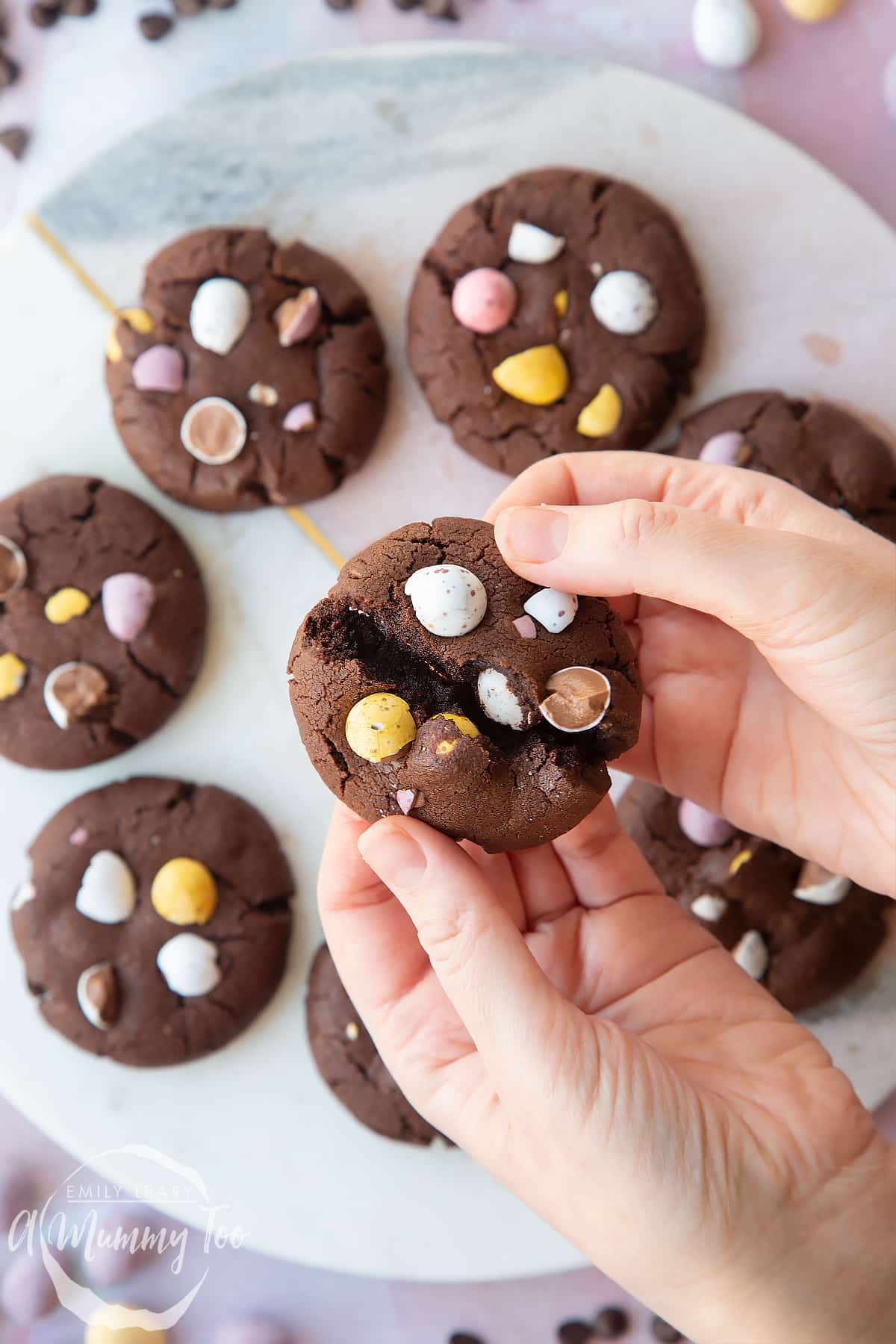 Hands breaking open a chocolate Easter cookie, with more arranged on a round marble board. The cookies are chocolate brown and topped with Mini Eggs.