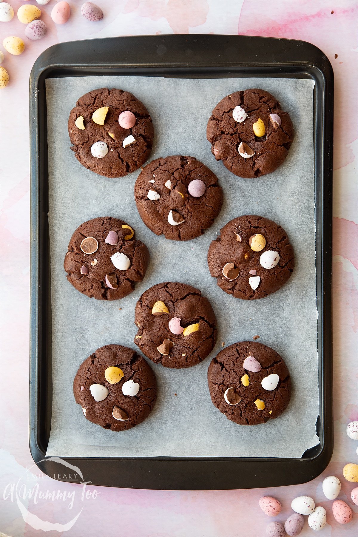 Chocolate Easter cookies arranged on a baking tray. The cookies are chocolate brown and topped with Mini Eggs. 