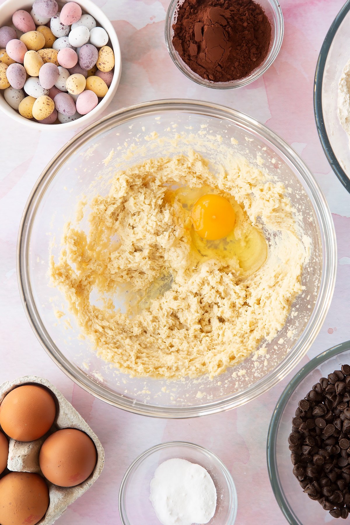 Butter and golden granulated sugar beaten together in a bowl with an egg on top. Ingredients to make Chocolate Easter cookies surround the bowl.