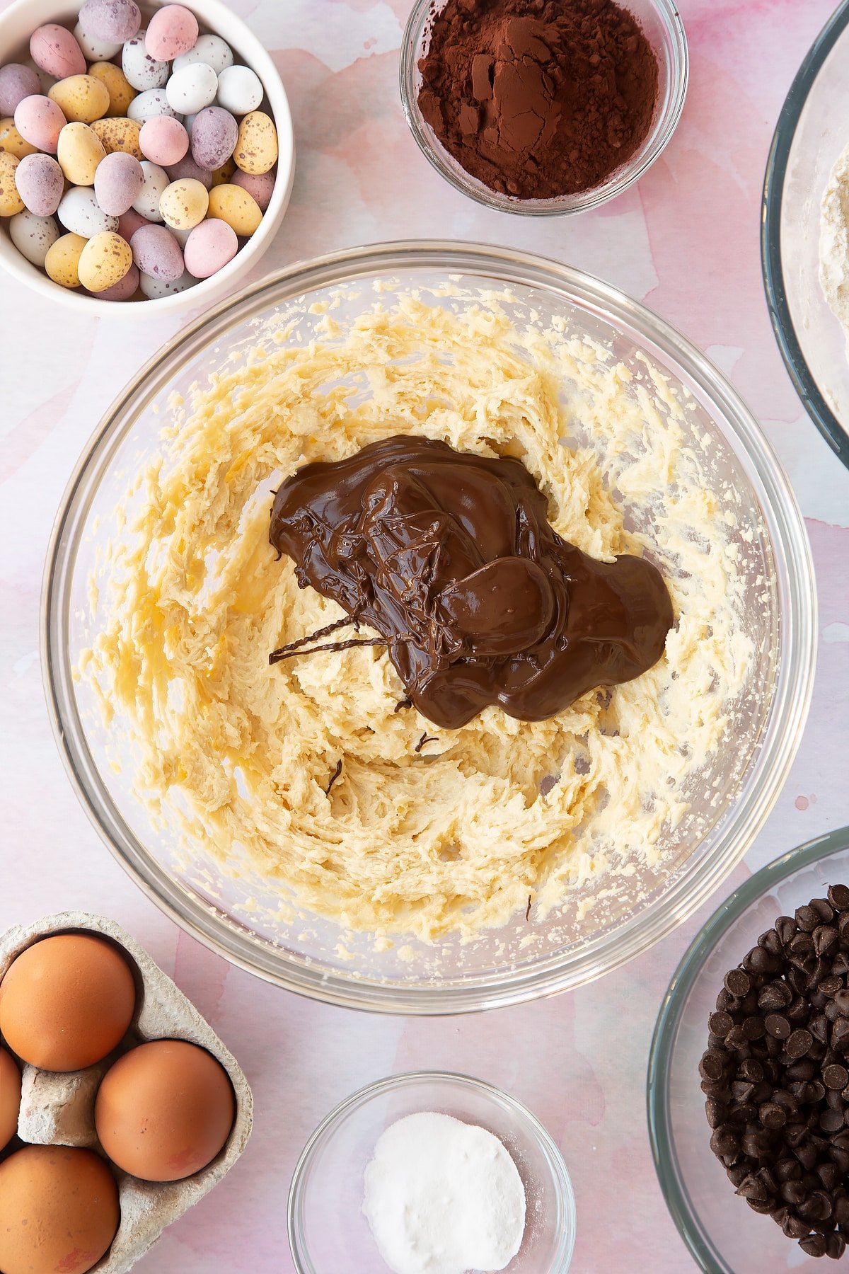 Butter, golden granulated sugar and egg beaten together in a bowl with melted dark chocolate on top. Ingredients to make Chocolate Easter cookies surround the bowl.