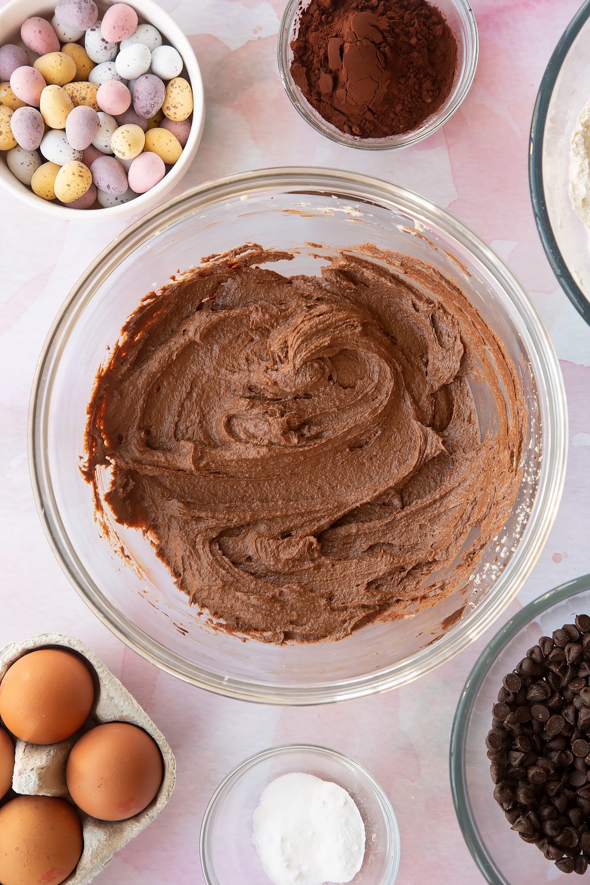 Butter, golden granulated sugar, egg and dark chocolate beaten together in a bowl. Ingredients to make Chocolate Easter cookies surround the bowl.