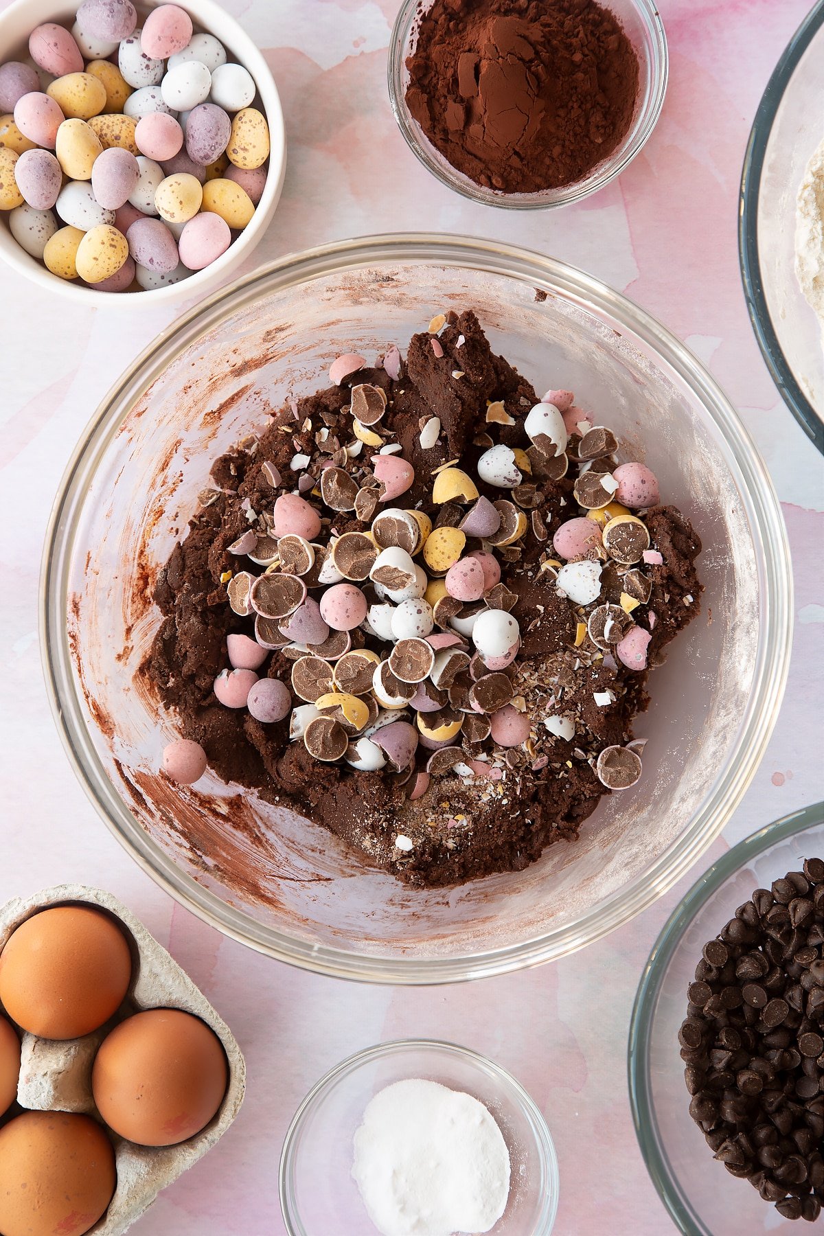 Chocolate cookie dough in a bowl with smashed Mini Eggs on top. Ingredients to make Chocolate Easter cookies surround the bowl.