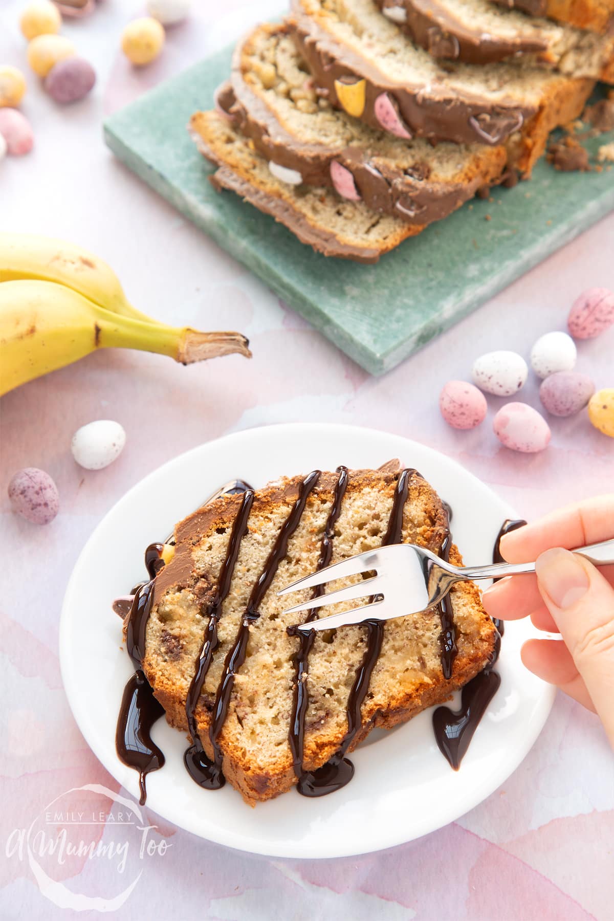 A slice of Easter banana bread on a white plate. The slice is drizzled with chocolate sauce and a hand holding a fork digs into it.