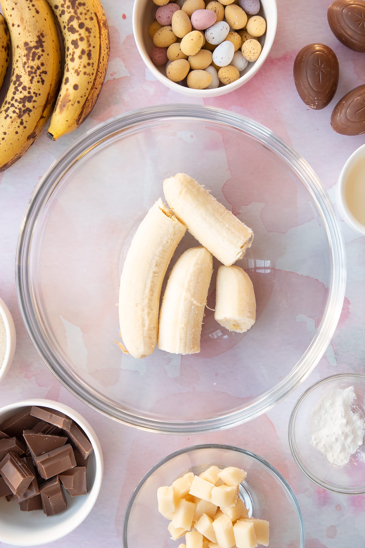 Bananas in a mixing bowl. Ingredients to make Easter banana bread surround the bowl.