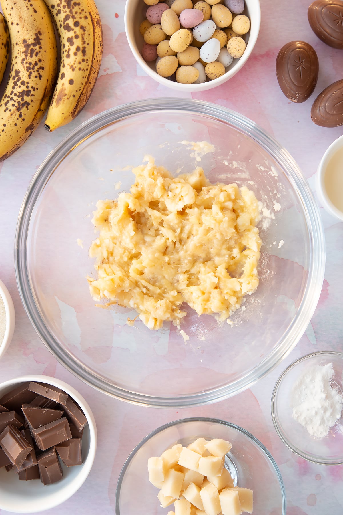 Mashed bananas in a mixing bowl. Ingredients to make Easter banana bread surround the bowl.