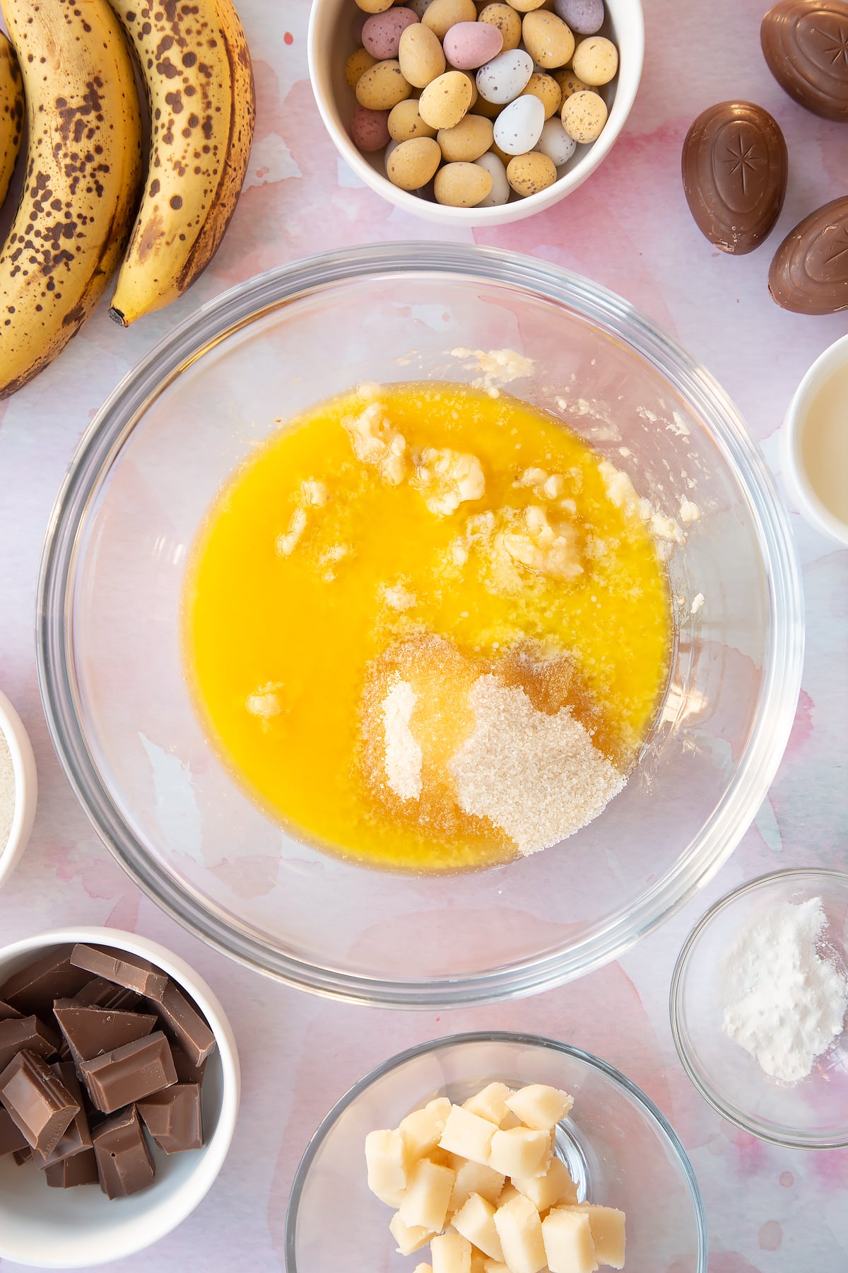 Mashed bananas topped with melted butter and sugar in a mixing bowl. Ingredients to make Easter banana bread surround the bowl.