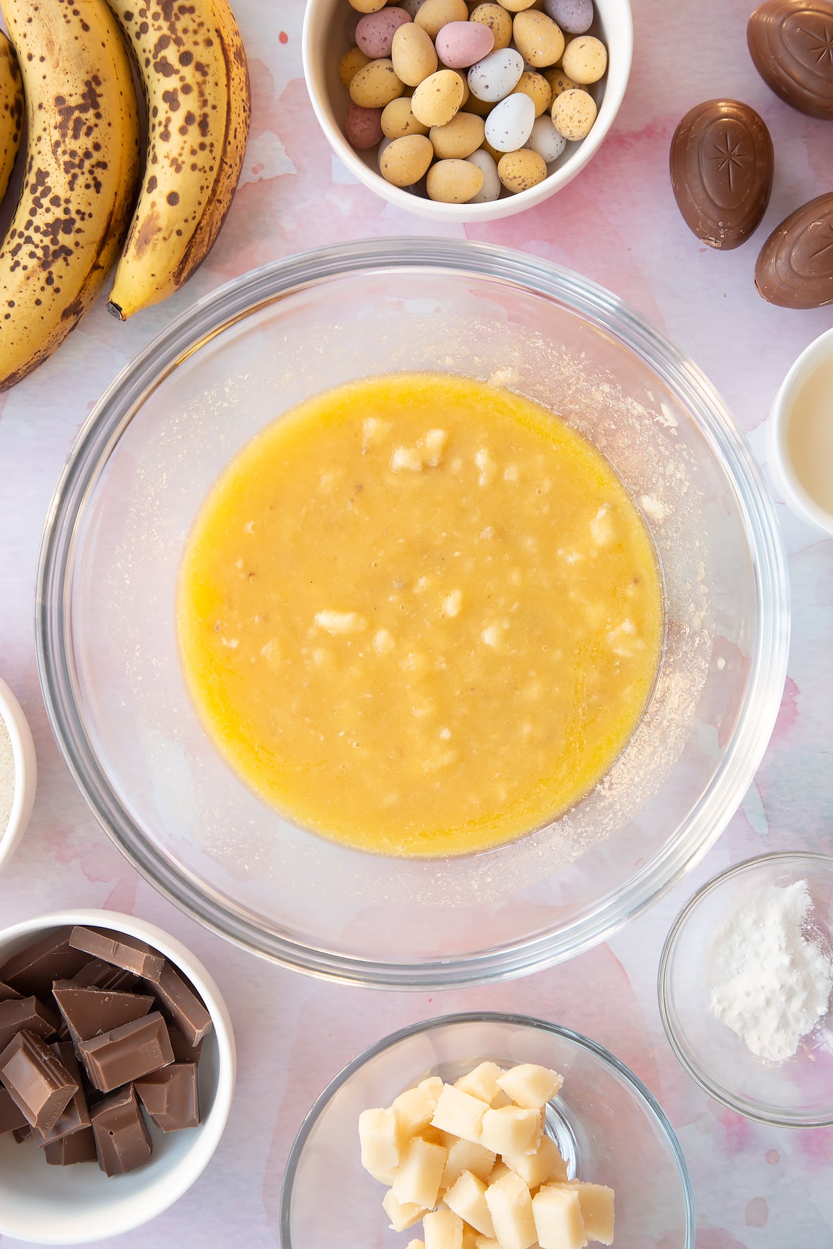 Mashed bananas, melted butter and sugar in a mixing bowl. Ingredients to make Easter banana bread surround the bowl.