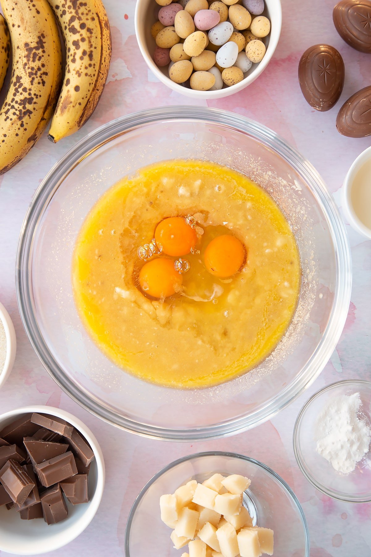 Mashed bananas, melted butter and sugar in a mixing bowl with eggs on top. Ingredients to make Easter banana bread surround the bowl.