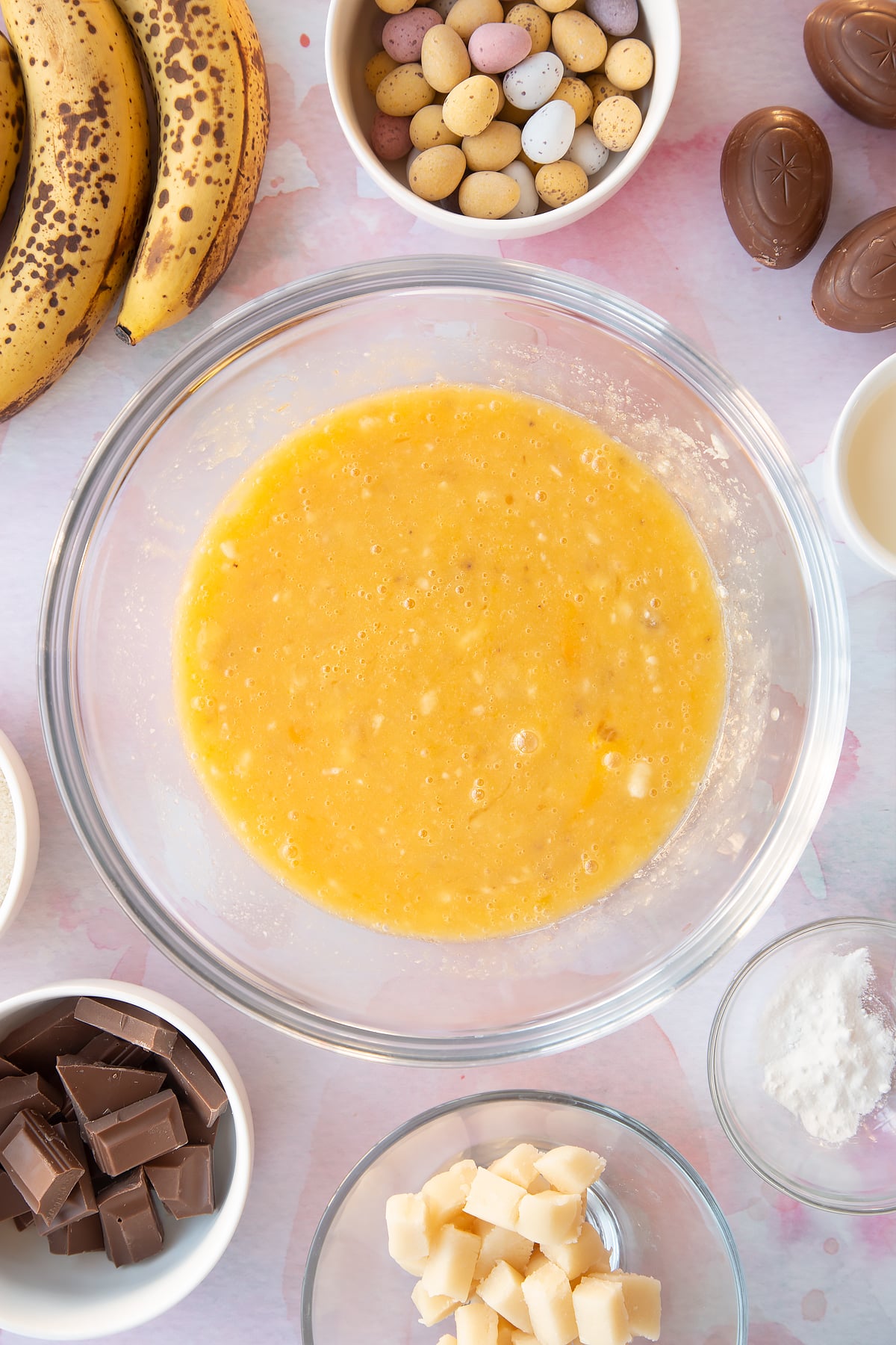 Mashed bananas, melted butter, sugar and eggs in a mixing bowl. Ingredients to make Easter banana bread surround the bowl.