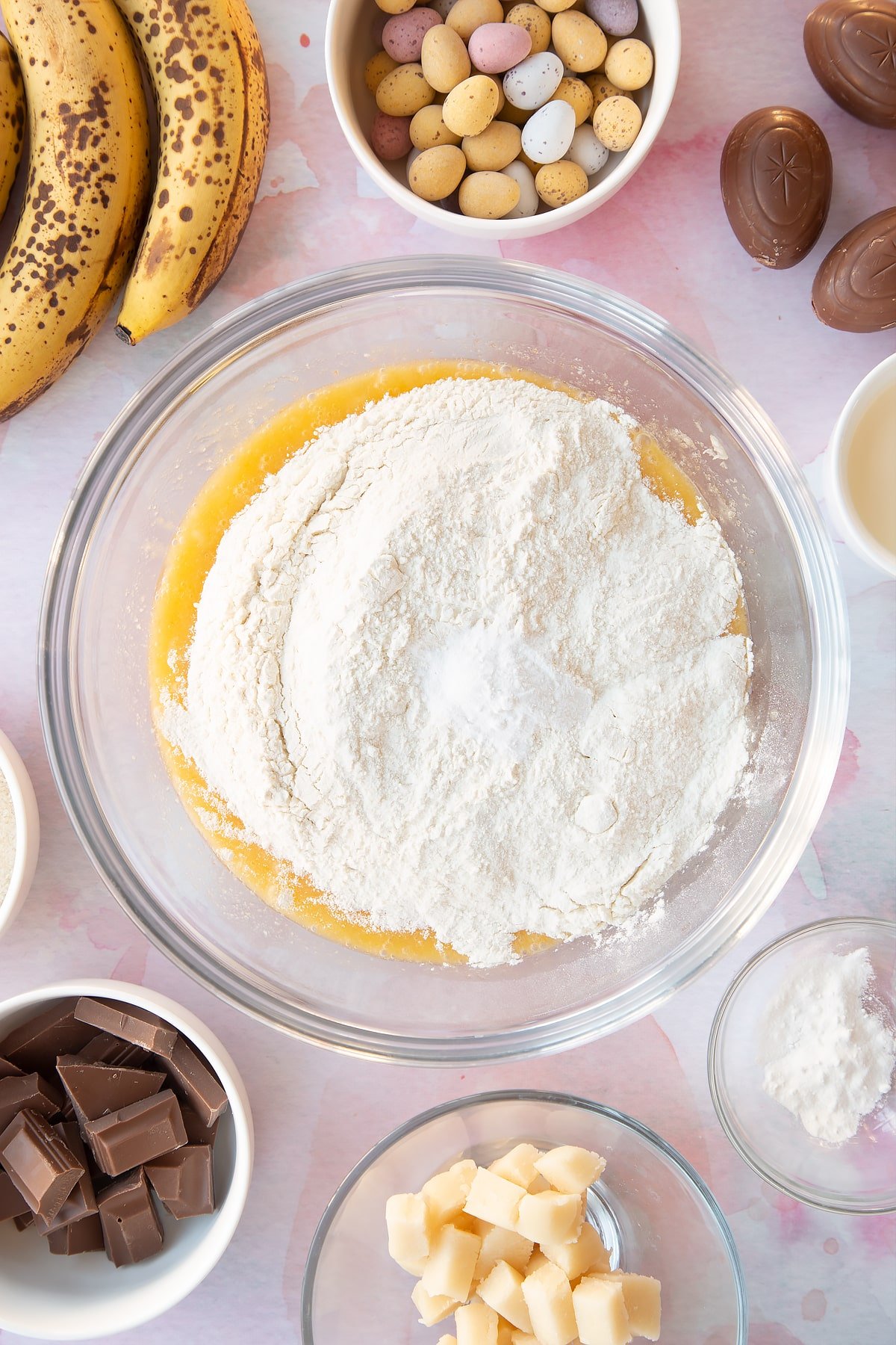 Mashed bananas, melted butter, sugar and eggs in a mixing bowl with flour and baking powder on top. Ingredients to make Easter banana bread surround the bowl.