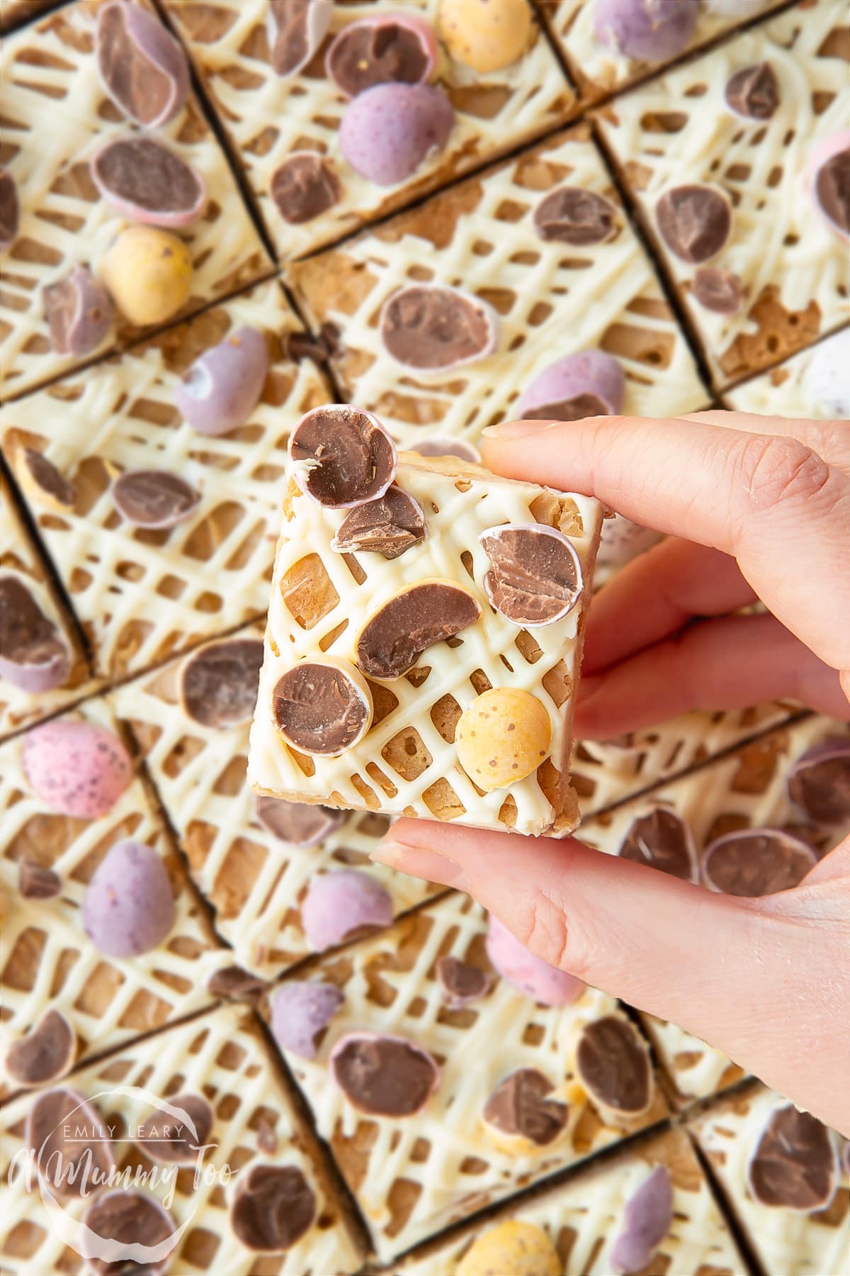 Hand holding a piece of Mini Egg blondie. More Mini Egg blondies are in the background.