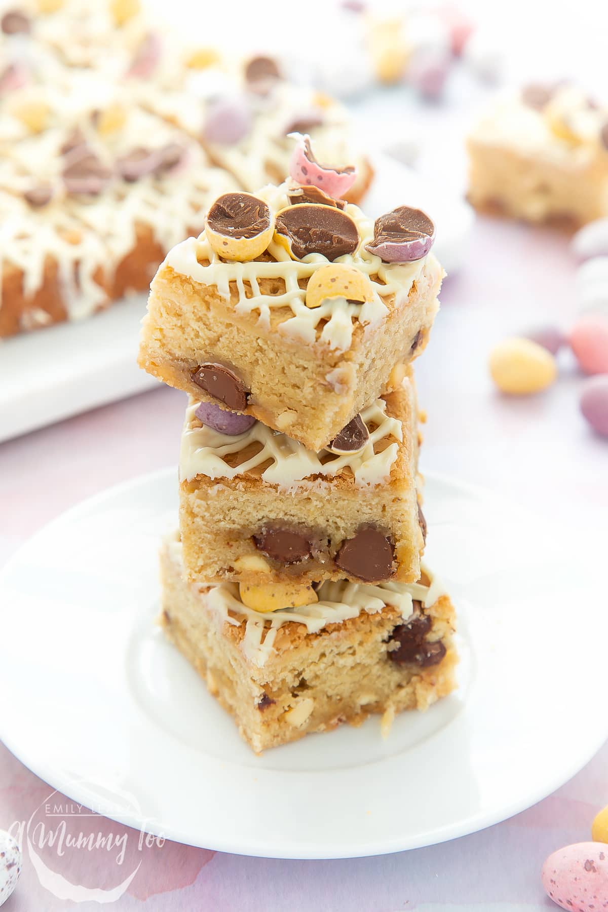 Three Mini Egg blondies on a white place. More Mini Egg blondies are on a white behind in the background.