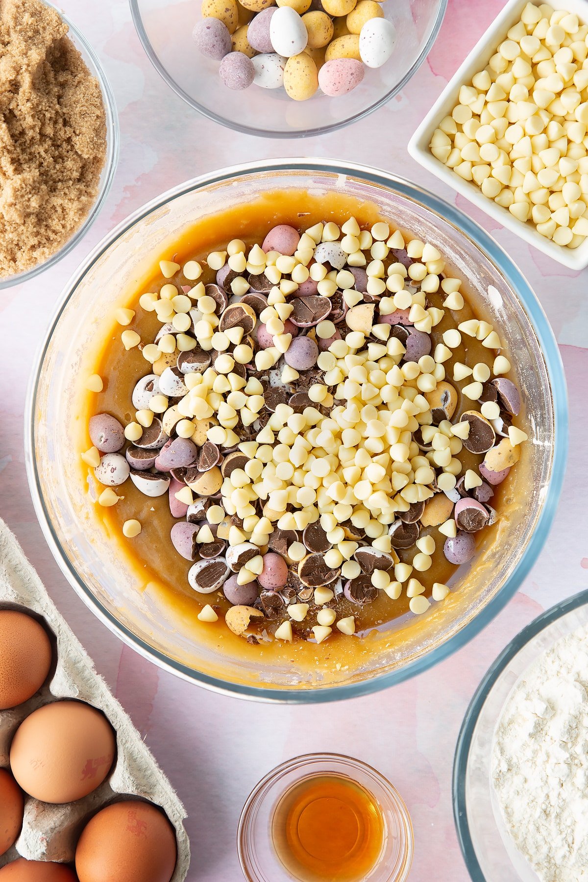 Basic blondie batter in a bowl with white choc chips and mini eggs on top. Ingredients to make Mini Egg blondies surround the bowl. 