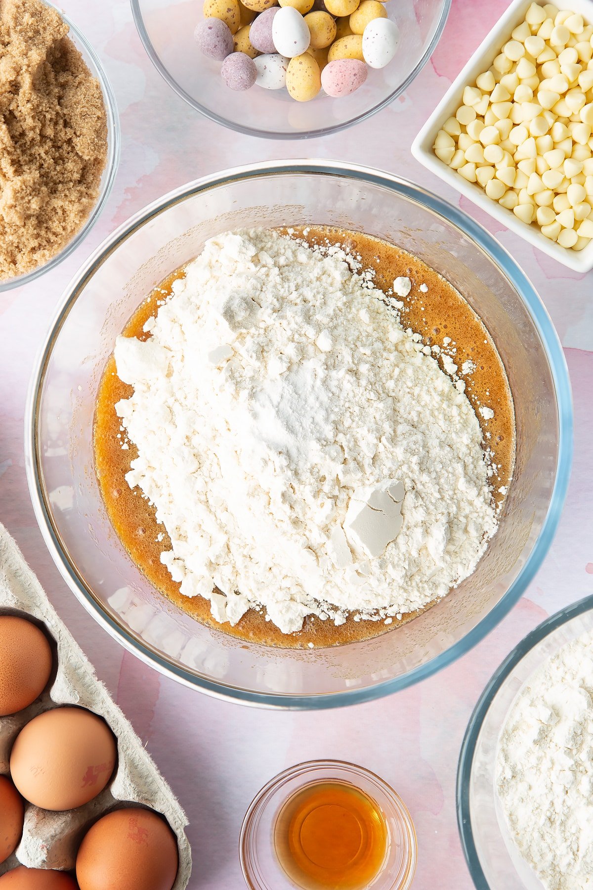 Melted butter, sugar and eggs mixed together in a bowl with flour & baking powder on top. Ingredients to make Mini Egg blondies surround the bowl. 