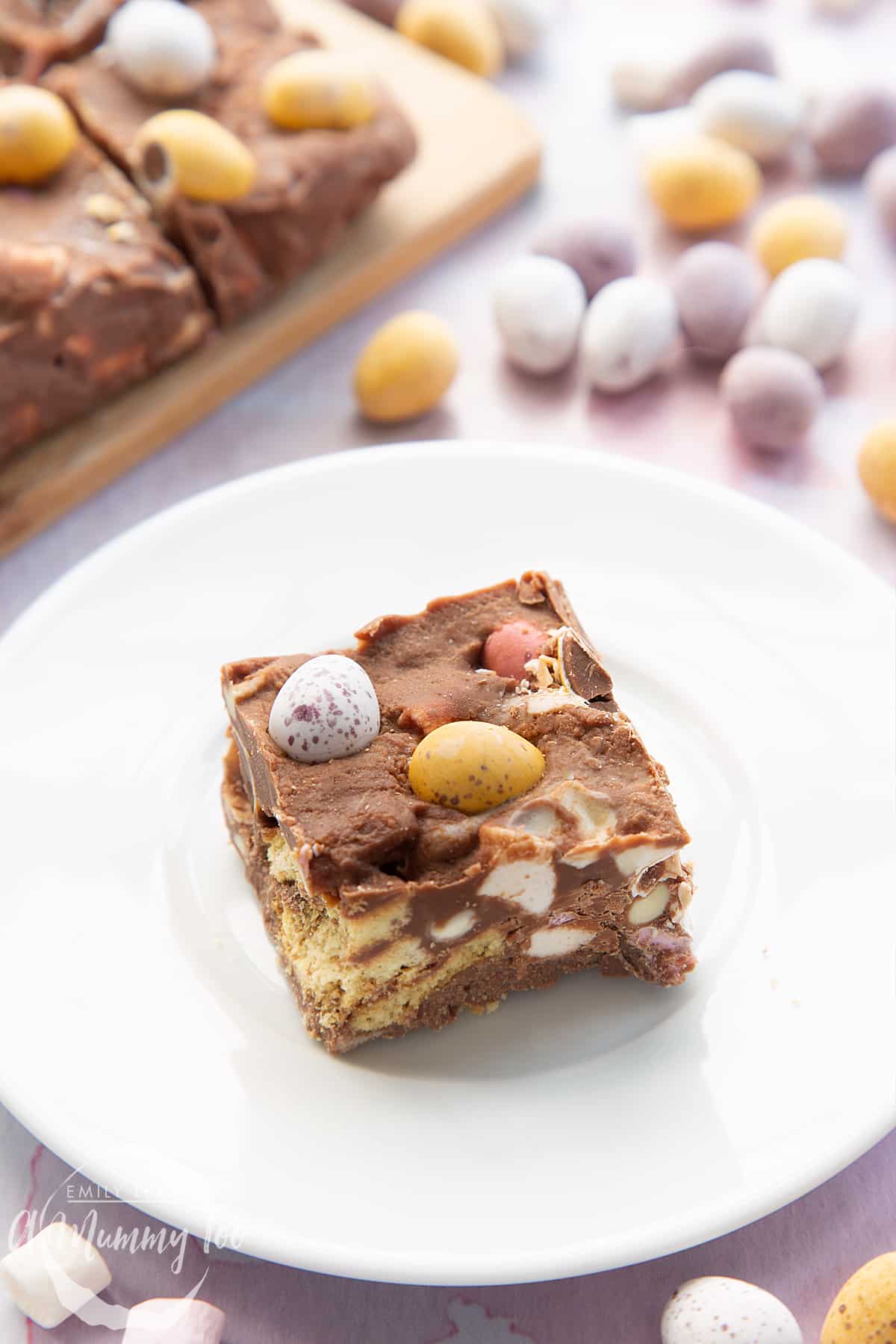 A piece of Mini Egg rocky road on a small white plate. More rocky road is in the background.