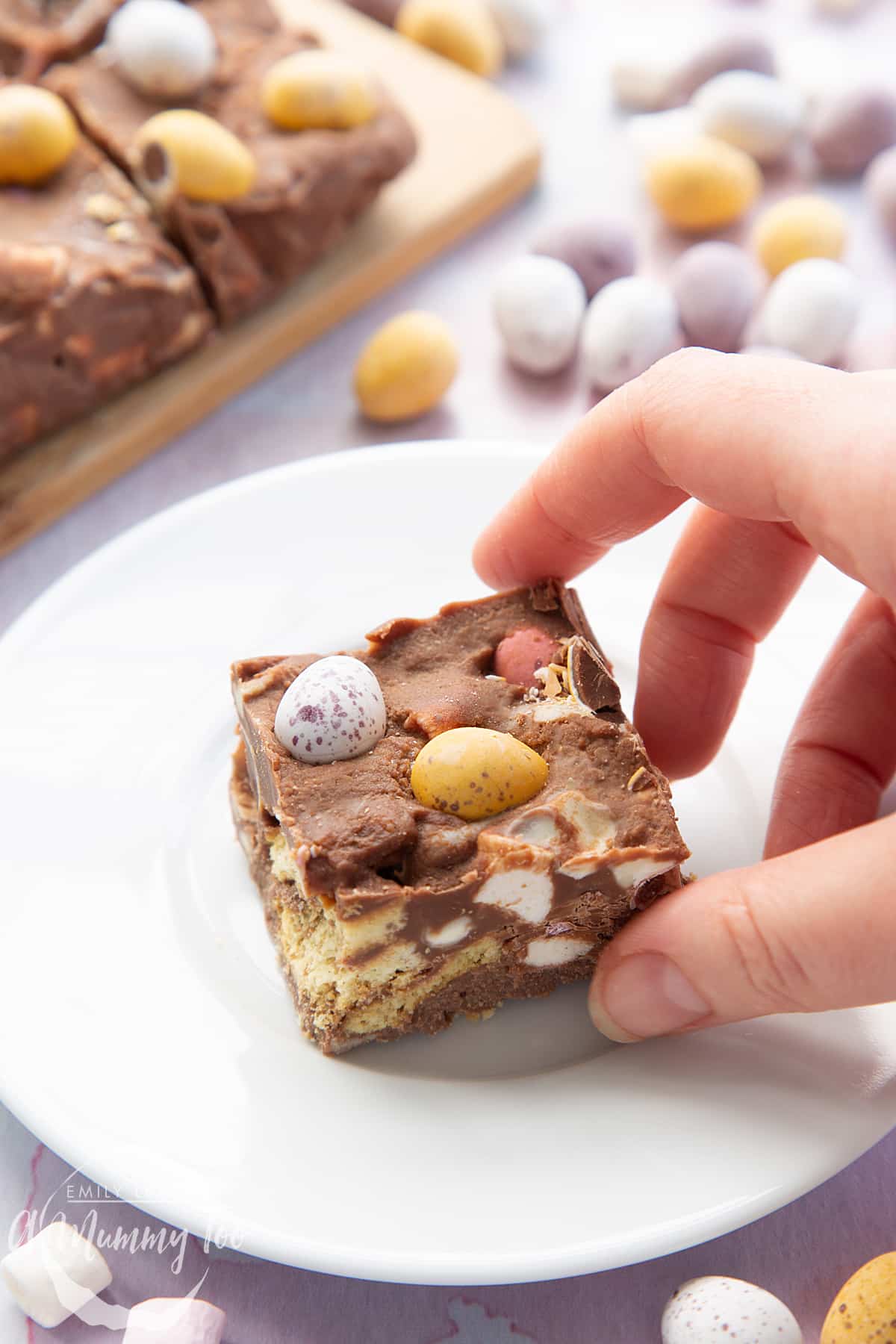 A piece of Mini Egg rocky road on a small white plate. A hand reaches to take it.