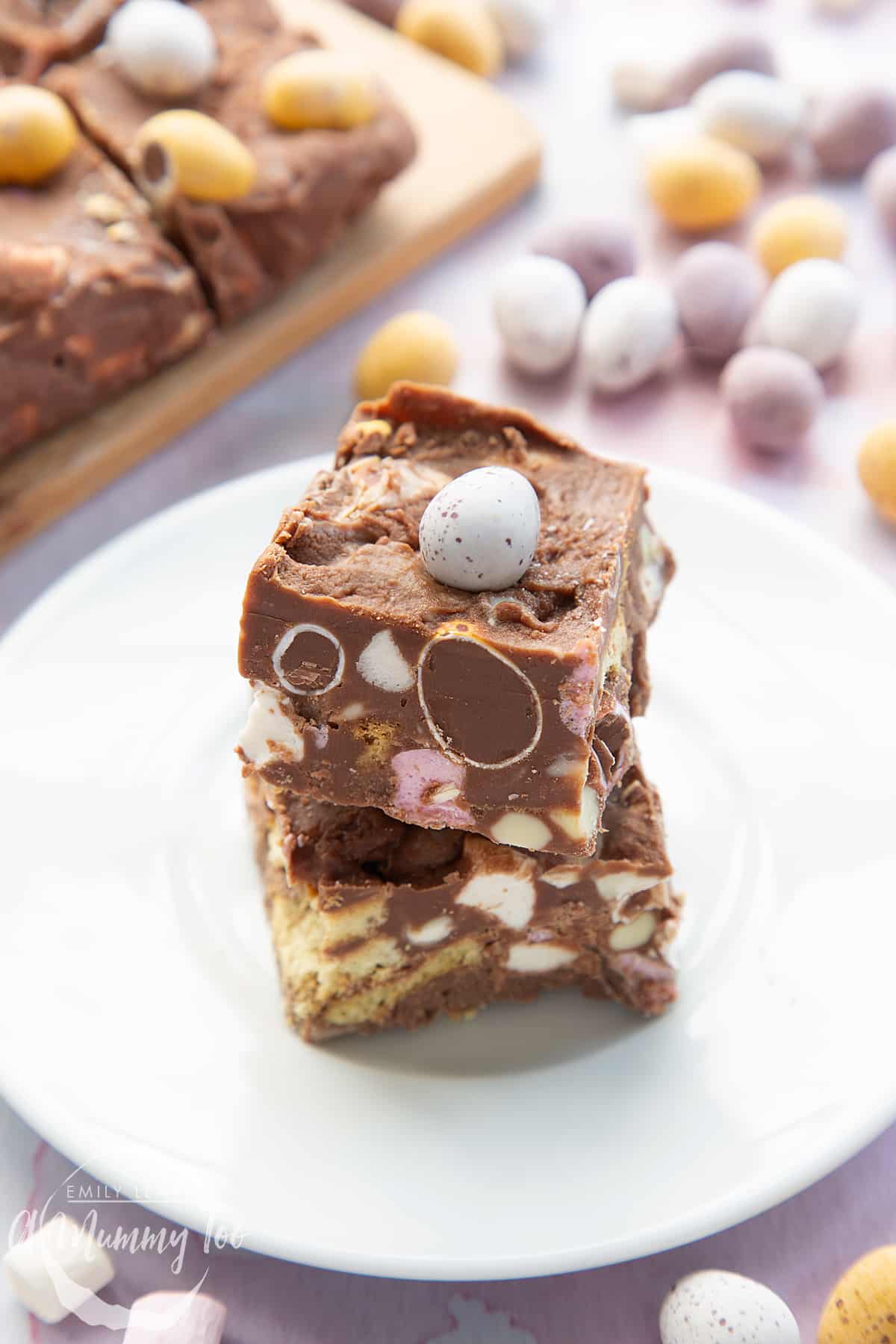 Two squares of Mini Egg rocky road stacked on a small white plate. Mini Eggs surround the plate.