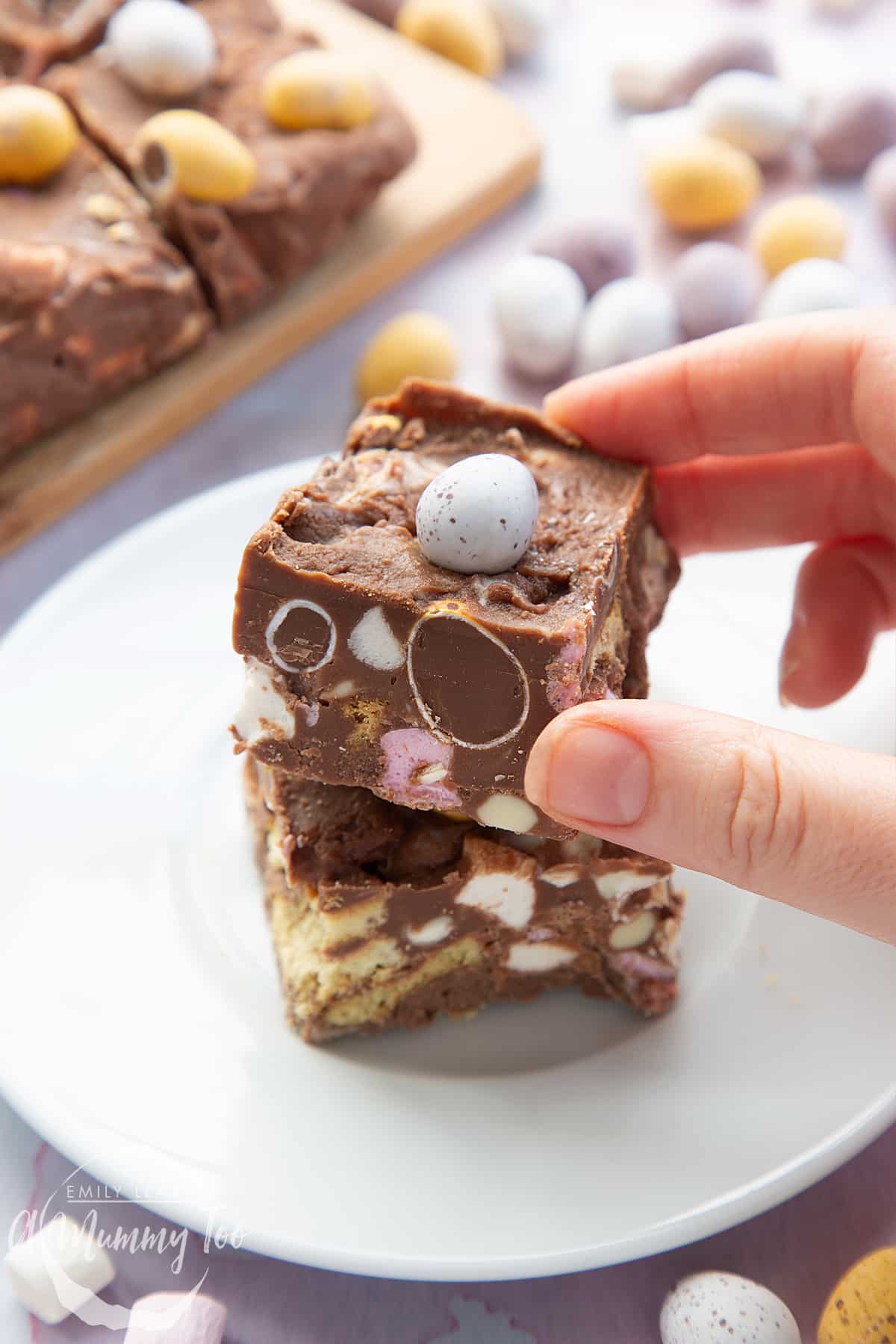 Two squares of Mini Egg rocky road stacked on a small white plate. A hand reaches to take the top piece.