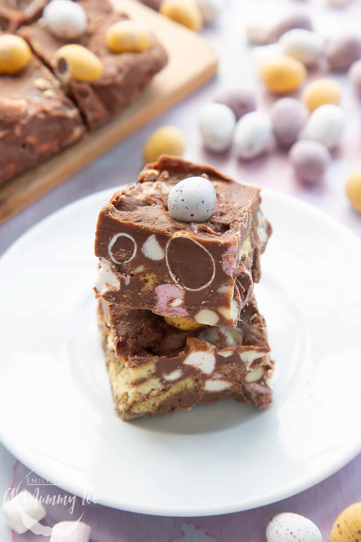 Two squares of Mini Egg rocky road stacked on a small white plate. More rocky road is in the background.